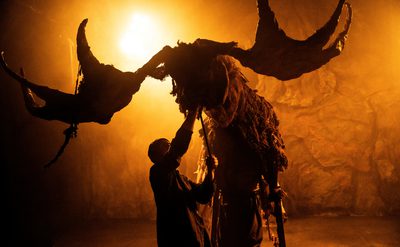 A large Elk puppet, operated by three people, is lit in golden light, shining from behind.