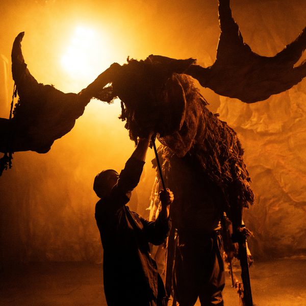 A large Elk puppet, operated by three people, is lit in golden light, shining from behind.